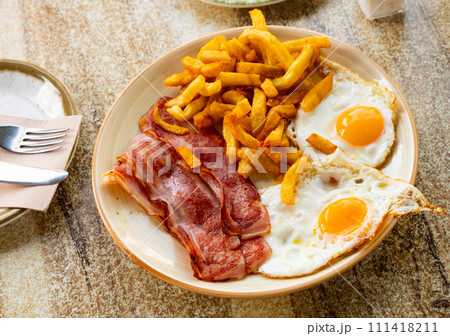 Plate of appetizing hearty breakfast of fried eggs with slices of delicious bacon and French fries Plate of appetizing hearty breakfast of fried eggs with slices of delicious bacon and French fries 111418211