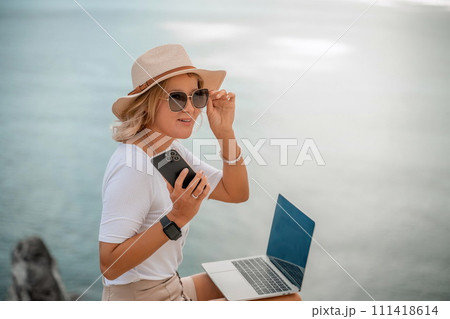 Freelance women sea working on the computer. Good looking middle aged woman typing on a laptop keyboard outdoors with a beautiful sea view. The concept of remote work. Freelance women sea working on the computer. Good looking middle aged woman typing on a laptop keyboard outdoors with a beautiful sea view. The concept of remote work. 111418614