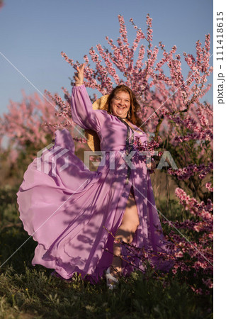 Woman blooming peach orchard. Against the backdrop of a picturesque peach orchard, a woman in a long pink dress and hat enjoys a peaceful walk in the park, surrounded by the beauty of nature. Woman blooming peach orchard. Against the backdrop of a picturesque peach orchard, a woman in a long pink dress and hat enjoys a peaceful walk in the park, surrounded by the beauty of nature. 111418615