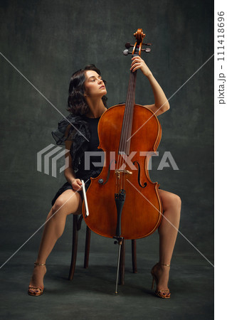 Elegant, beautiful young woman, cellist sitting with wooden cello against dark green vintage background. Acoustic concert Elegant, beautiful young woman, cellist sitting with wooden cello against dark green vintage background. Acoustic concert 111418696