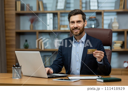 Confident mature businessman in suit with credit card, using laptop at messy home office desk, symbolizing remote work. 111418929