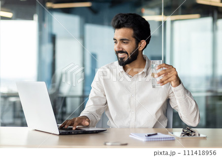 A white-collar worker with a beard is multitasking, drinking water and using a laptop computer on a desk. A white-collar worker with a beard is multitasking, drinking water and using a laptop computer on a desk. 111418956