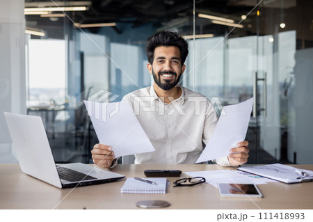 Indian businessman with a beard working at his desk with a laptop, reviewing papers in a modern office environment. Indian businessman with a beard working at his desk with a laptop, reviewing papers in a modern office environment. 111418993