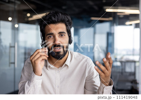 Confident young Indian entrepreneur working in office, talking through a headset with a focused expression. Confident young Indian entrepreneur working in office, talking through a headset with a focused expression. 111419048