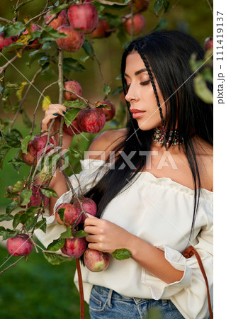 Sad, dark-haired woman in shoulderless white shirt, holding tree branches, laden with red apples. Close up of thoughtful pierced girl posing in apple orchard with closed eyes. Harvest, beauty concept. Sad, dark-haired woman in shoulderless white shirt, holding tree branches, laden with red apples. Close up of thoughtful pierced girl posing in apple orchard with closed eyes. Harvest, beauty concept. 111419137