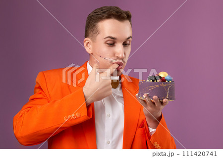 handsome birthday man in orange jacket eating cake on purple background handsome birthday man in orange jacket eating cake on purple background 111420741