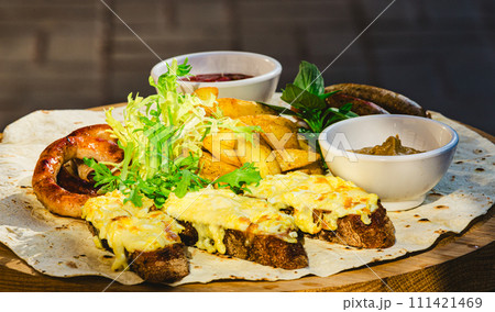 Grilled sausages with potato wedges and bread with cheese and sauces close-up on a wooden tray. 111421469