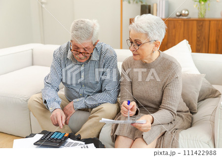 Middle aged senior couple sit with laptop and paper document. Older mature man woman reading paper bill pay online at home managing bank finances calculating taxes planning loan debt pension payment Middle aged senior couple sit with laptop and paper document. Older mature man woman reading paper bill pay online at home managing bank finances calculating taxes planning loan debt pension payment 111422988