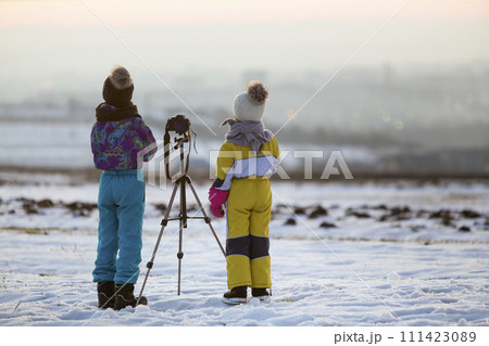 Two children boy and girl having fun outside in winter playing with photo camera on a tripod on snow covered field. 111423089