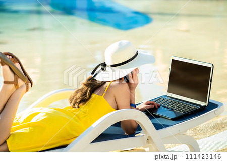 Young woman in yellow dress is laying on beach chair working on computer laptop connected to wireless internet typing text on keys in summer resort. Doing studies while travelling concept. Young woman in yellow dress is laying on beach chair working on computer laptop connected to wireless internet typing text on keys in summer resort. Doing studies while travelling concept. 111423146