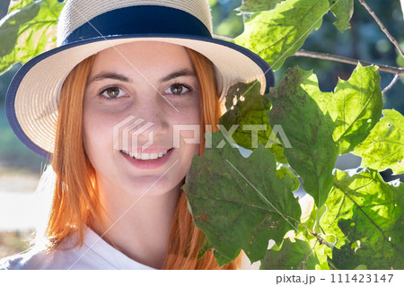 Portrait of pretty young teenage girl with red hair in yellow hat near green tree leaves. 111423147