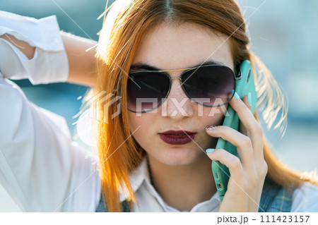 Closeup portrait of fashionable young redhead woman in sunglasses talking on cell phone outdoors in summer. Closeup portrait of fashionable young redhead woman in sunglasses talking on cell phone outdoors in summer. 111423157