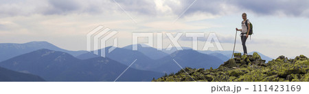 Young slim blond tourist girl with backpack and stick standing on rocky top against bright blue morning sky on foggy mountain range panorama background. Tourism, traveling and climbing concept. 111423169