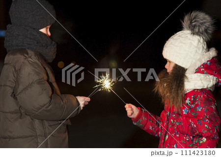 Profile of two cute young children, boy and girl in warm winter clothing holding burning sparkler fireworks on dark night outdoors copy space background. New Year and Christmas celebration concept. Profile of two cute young children, boy and girl in warm winter clothing holding burning sparkler fireworks on dark night outdoors copy space background. New Year and Christmas celebration concept. 111423180