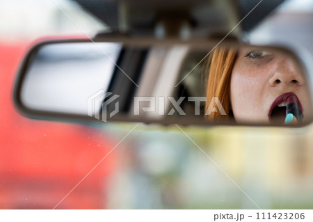 Closeup of a young redhead woman driver correcting her makeup with dark red lipstick looking in car rearview mirror behind steering wheel of a vehicle. 111423206