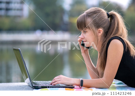 Young female office employee working behind laptop computer while talking on cellphone lying down in summer park outdoors. Remote work concept. 111423246