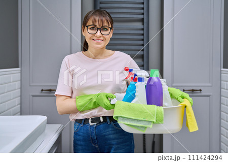 Middle-aged woman in gloves with basin of detergents in bathroom Middle-aged woman in gloves with basin of detergents in bathroom 111424294