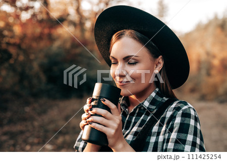 Hipster woman enjoys aroma and taste of fresh coffee. Drinking from thermos in autumn forest 111425254