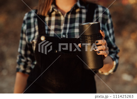 Hipster woman enjoys aroma and taste of fresh coffee. Drinking thermos in forest 111425263