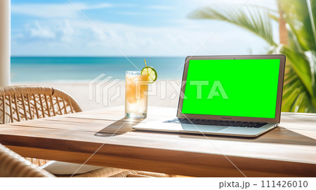 Laptop with green screen on a table by the beach with a tropical cocktail, palm trees, and ocean view portraying a relaxed work environment 111426010