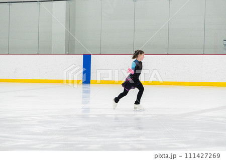 Young girl perfecting her figure skating routine while wearing her competition dress at an indoor ice rink. Young girl perfecting her figure skating routine while wearing her competition dress at an indoor ice rink. 111427269
