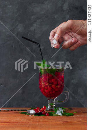 Summer red currant tea in a glass on dark background.Male hand holds melting ice cube with drop over fruit cold cocktail Summer red currant tea in a glass on dark background.Male hand holds melting ice cube with drop over fruit cold cocktail 111427436