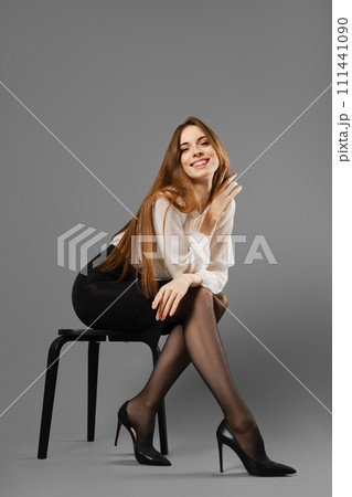 Smiling woman in pencil skirt and blouse leaning forward while sitting on a chair Smiling woman in pencil skirt and blouse leaning forward while sitting on a chair 111441090