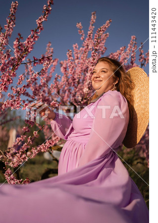 Woman blooming peach orchard. Against the backdrop of a picturesque peach orchard, a woman in a long pink dress and hat enjoys a peaceful walk in the park, surrounded by the beauty of nature. 111443420