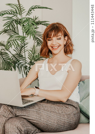 Brunette macbook sofa. Attractive young woman working on a laptop while sitting on the couch at home. She is wearing a light blouse and trousers. 111443448