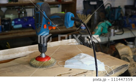 a drill with an attachment made of metal bristles standing on a wooden board in a craftsman's workshop, an apparatus for working with wood and a dust mask as a carpenter's working tools on a workbench a drill with an attachment made of metal bristles standing on a wooden board in a craftsman's workshop, an apparatus for working with wood and a dust mask as a carpenter's working tools on a workbench 111447359