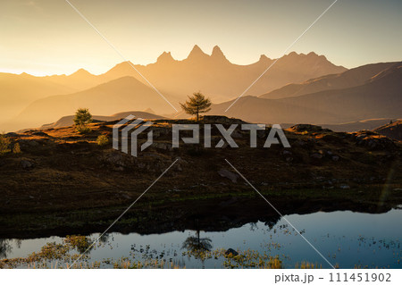 Golden sunrise over Arves massif with lonely tree relfection on Lac Guichard at French Alps Golden sunrise over Arves massif with lonely tree relfection on Lac Guichard at French Alps 111451902