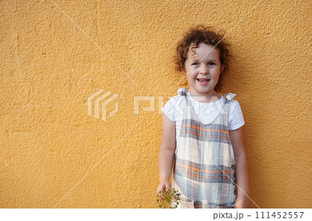 Happy cute kid child girl with curly hair, girl in stylish overalls holding a plant, orange color background with copy space 111452557