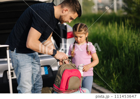 Dad and daughter are standing with their suitcases near a car with an open trunk. Happy little girl preparing to travel. 111453647