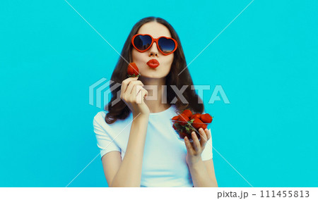 Portrait of woman with handful of fresh strawberries blowing lips in red heart shaped sunglasses 111455813
