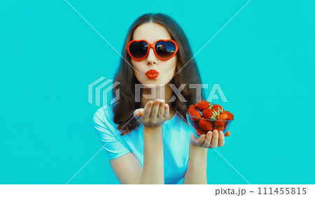 Portrait of woman with handful of fresh strawberries blowing lips in red heart shaped sunglasses Portrait of woman with handful of fresh strawberries blowing lips in red heart shaped sunglasses 111455815