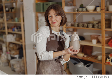 Portrait of young female potter in apron with mug looking at camera while posing in workshop Portrait of young female potter in apron with mug looking at camera while posing in workshop 111456194