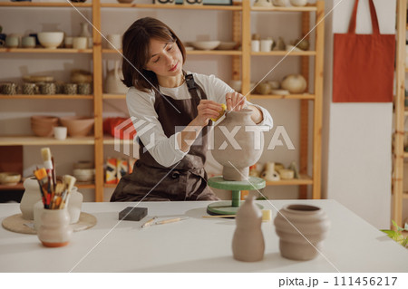 Smiling young female ceramist wearing apron work with unfired clay vase in pottery studio Smiling young female ceramist wearing apron work with unfired clay vase in pottery studio 111456217