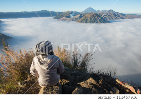 Back view of women sitting on the hill and looking to the beautiful sunrise and sea of fog over Bromo Tengger Semeru National Park in East Java, Indonesia. Back view of women sitting on the hill and looking to the beautiful sunrise and sea of fog over Bromo Tengger Semeru National Park in East Java, Indonesia. 111457916