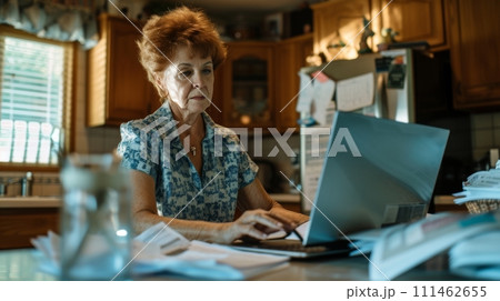 Old woman doing taxes on laptop in a kitchen with document on table 111462655