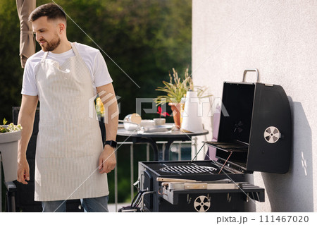 Side view of handsome man try how hot is BBQ grill. Man in apron make dinner outdoor 111467020