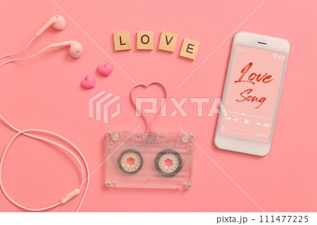 vintage transparent audio cassette magnetic tape in shape of heart, earphones, mobile phone playing love song on screen. flat lay Valentine's Day music on pink background 111477225