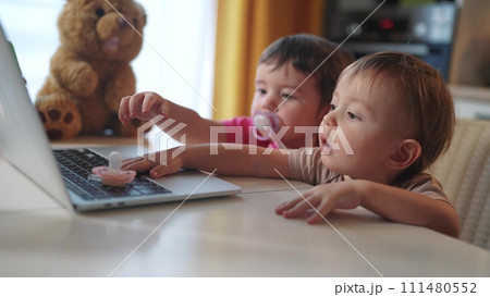 twin baby children a playing laptop watching video in the kitchen. happy family kid dream concept. baby twins playing video game lifestyle on laptop looking at screen in kitchen 111480552