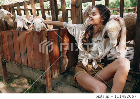 A woman is sitting in front of a fence with sheep in a pen with her hand on the fence 111482067