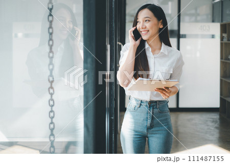 Buiness technology concept, Young businesswoman in a white shirt, engaging in a phone conversation while holding a clipboard in a sunlit modern office. Buiness technology concept, Young businesswoman in a white shirt, engaging in a phone conversation while holding a clipboard in a sunlit modern office. 111487155
