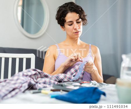 Young woman repairing plaid shirt with needle and thread Young woman repairing plaid shirt with needle and thread 111491034