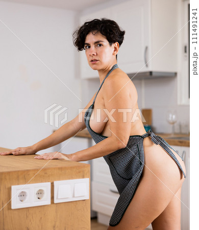 Sensual Latin woman in checkered apron dressed on naked corpus leaning kitchen table at flat 111491241