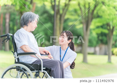 公園を散歩する車椅子の高齢者と介護士・看護師・理学療法士・運動指導員の女性 公園を散歩する車椅子の高齢者と介護士・看護師・理学療法士・運動指導員の女性 111492976