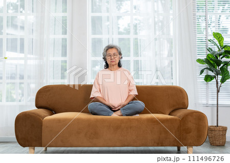 Mature Asian woman doing yoga for meditation on sofa in living room at home.Clam of senior or elderly women meditation deep breath and relax at cozy home.Happy Retired Meditation at home concept 111496726