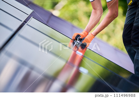 Worker building photovoltaic solar panel system on rooftop of house. Close up of man engineer in gloves installing solar module with help of hex key outdoors. Renewable energy generation concept. Worker building photovoltaic solar panel system on rooftop of house. Close up of man engineer in gloves installing solar module with help of hex key outdoors. Renewable energy generation concept. 111496988
