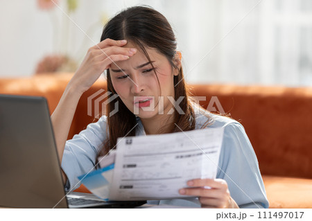Young Asian woman looks stressed while examining a bill, impactful financial pressure, anxiety, and the challenges of managing expenses. Financial problem. 111497017